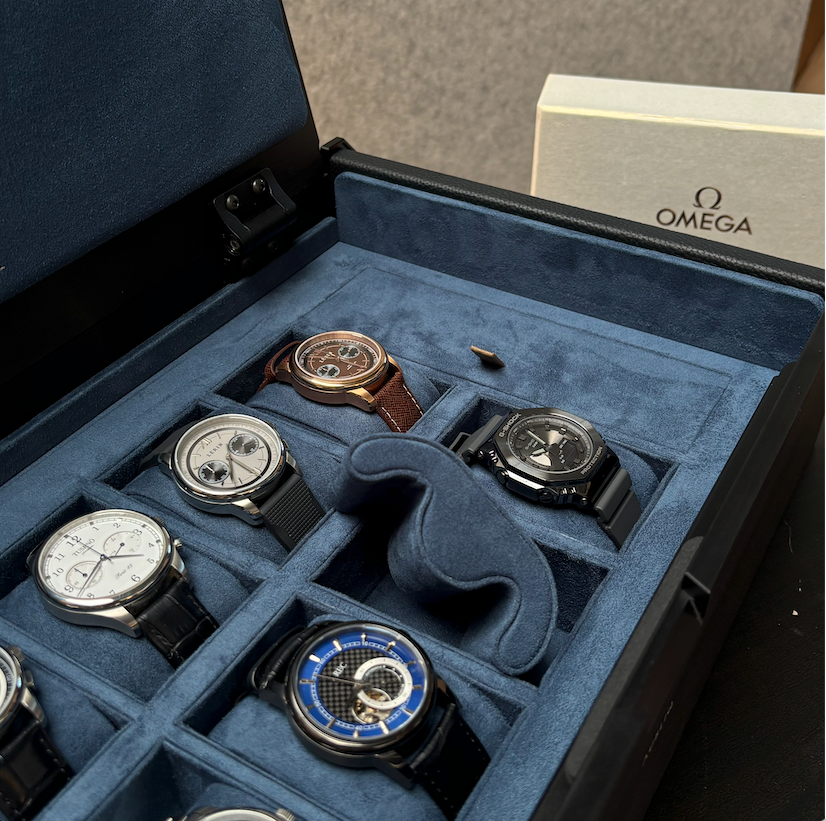A luxury watch display box with a blue velvet interior, showcasing several Omega wristwatches in individual slots. A white Omega-branded box is partially visible in the background.
