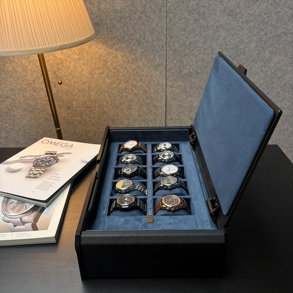 A black textured watch storage box with an open blue velvet interior holding ten watches, placed on a dark table next to a lamp and a magazine.