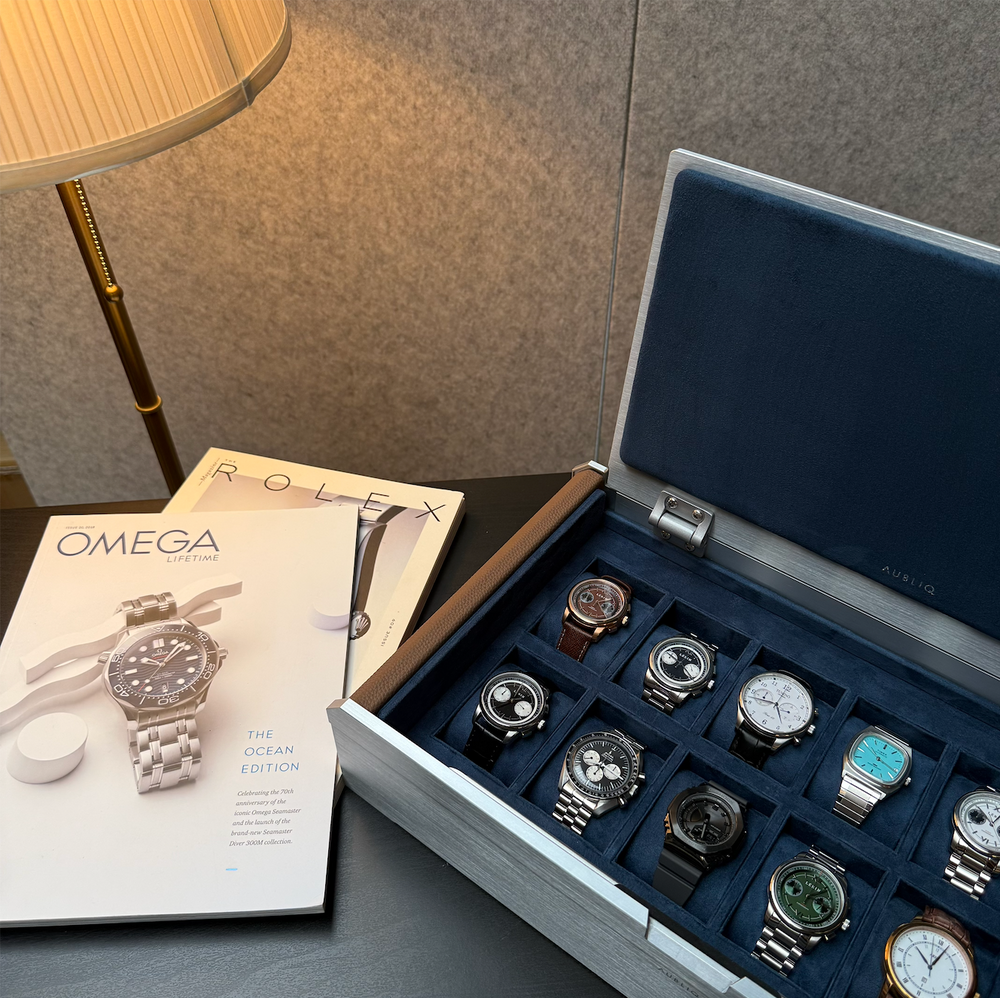 AUBLIQ silver watch box with watches in blue-lined compartments, displayed beside watch magazines and a table lamp.