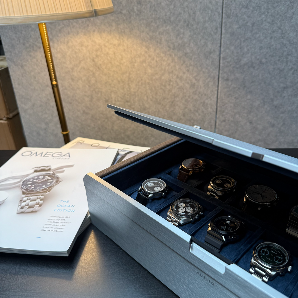 AUBLIQ silver watch box with its lid partly closed, revealing watches on blue cushions beside watch magazines.