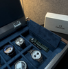 Close-up of the AUBLIQ silver watch box with its blue-lined strap compartment open, revealing a spare green watch strap.