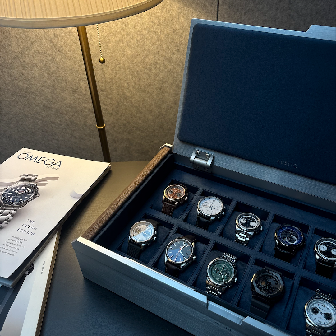 AUBLIQ silver watch box displaying watches in individual blue-lined compartments beside watch magazines and a table lamp.