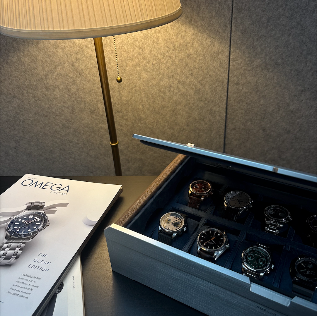 AUBLIQ silver watch box with its lid partly closed, revealing watches on blue cushions beside a table lamp.