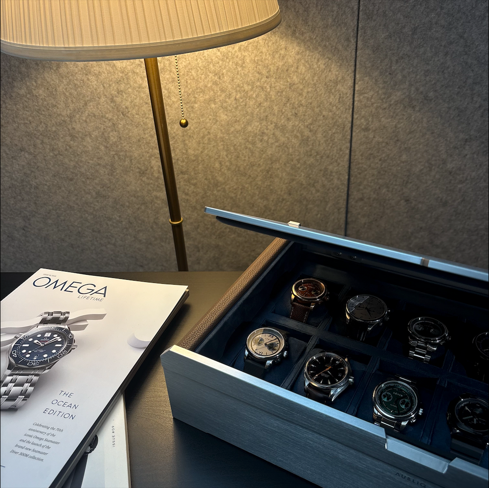 AUBLIQ silver watch box with its lid partly closed, revealing watches on blue cushions beside a table lamp.