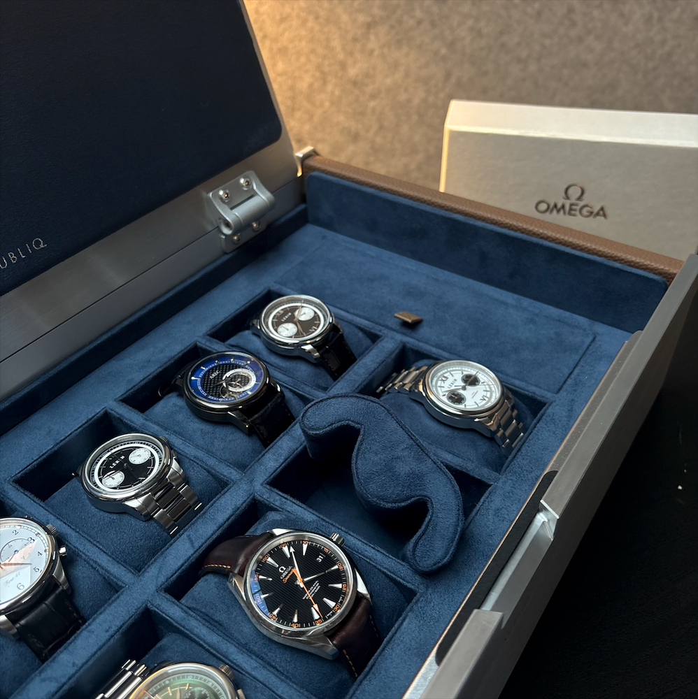 Close-up of the AUBLIQ silver watch box interior with a removed blue cushion, an empty watch slot and assorted watches.