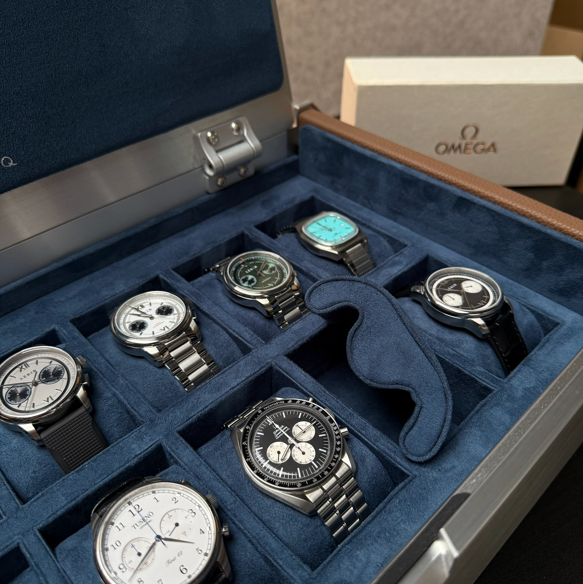 Close-up of the AUBLIQ silver watch box interior with assorted watches, blue-lined slots and a removed watch cushion.