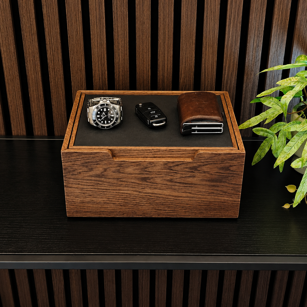 A rectangular wooden watch box with a dark interior tray holding a silver wristwatch, a black car key fob, and a brown leather wallet, displayed on a dark wooden shelf with a slatted wood wall background and a green plant at the side.